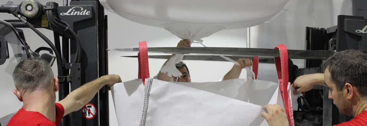 Workers opening the outlet spout of a suspended big bag at a discharging station